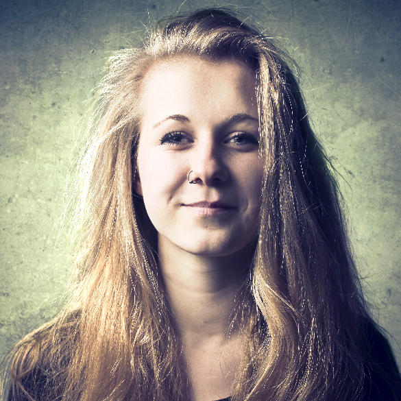A person with long, blonde hair and a nose piercing stands against a textured, grayish-green background. They are smiling slightly and are wearing a dark top.