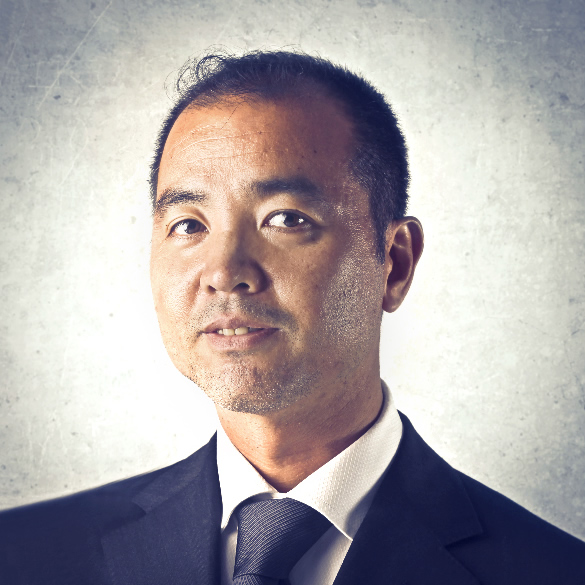 A man with short black hair and a light beard is wearing a dark suit, white dress shirt, and dark tie. He is looking slightly to the right. The background is a textured light gray color.